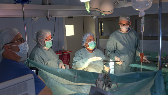 Dr. med. Susanne Born und Ihr Team stehen im OP-Saal und operieren einen Patienten. Der OTA reicht Dr. Susanne Born ein Operationswerkzeug. Das gesamte Team trägt grüne OP-Kleidung, Hauben und Mundschutz.
